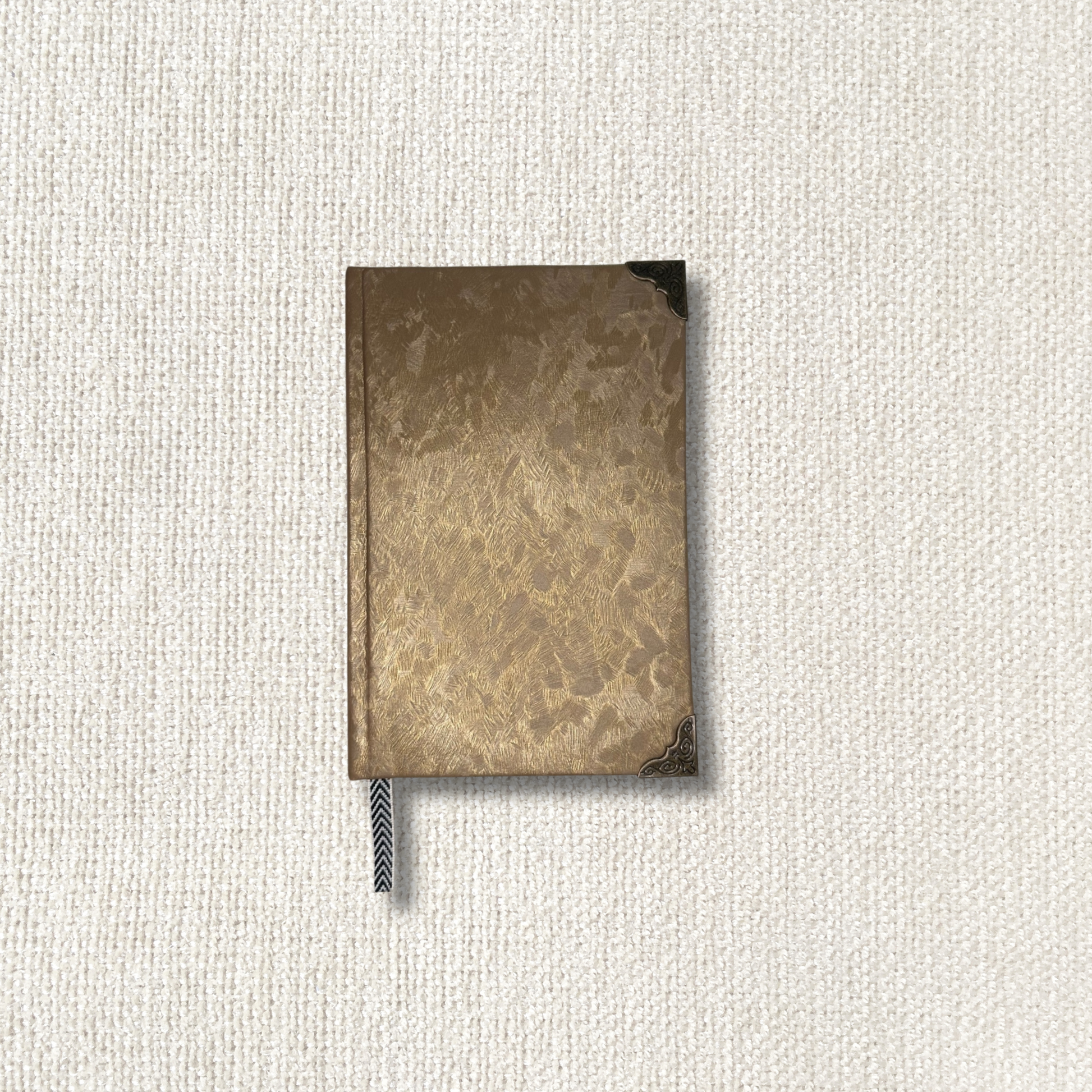 A small rectangular notebook or journal with a textured metallic gold cover and a dark ribbon bookmark, centered on a light beige woven fabric background.